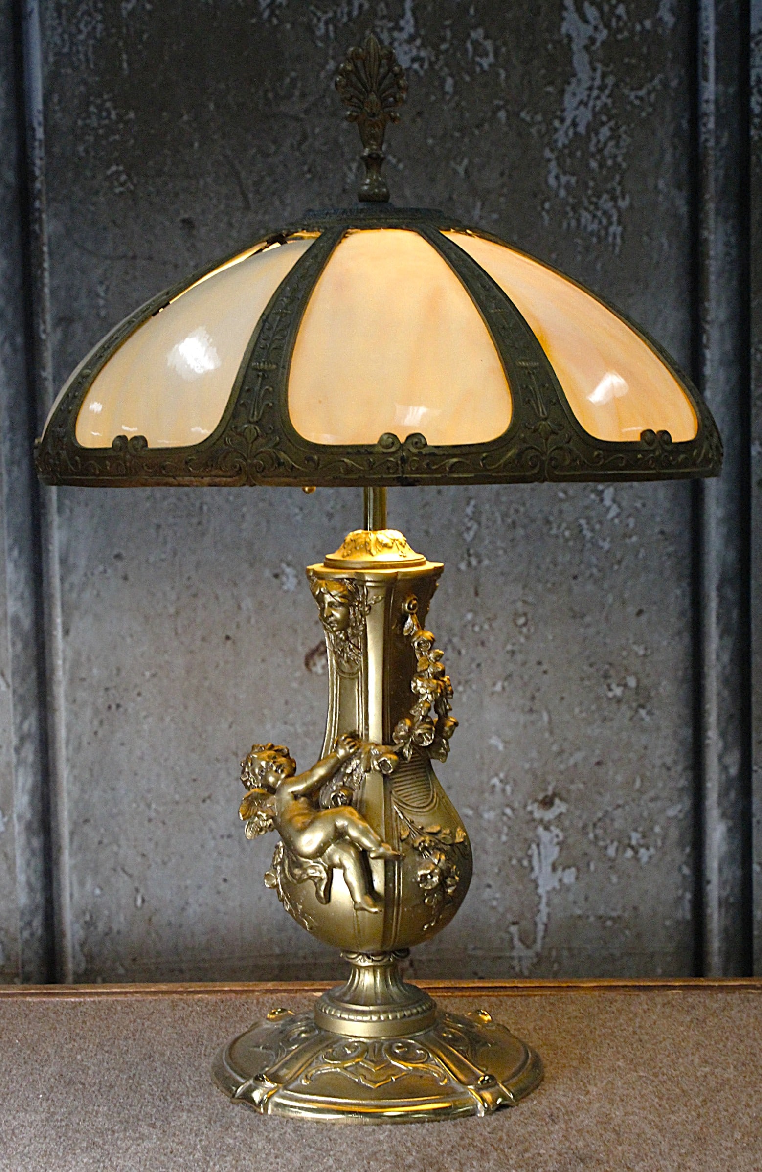 An Impressive Slag Panel Table Lamp: Early 20th century with figural base of a putto after L&F Moreau holding garland under a caramel slag bent panel shade. VG+ working cond, base refinished, shade original. Base embossed S2513, 23"H x 1