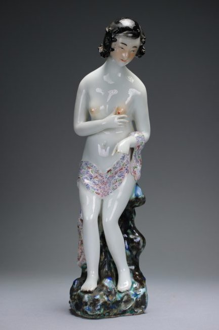 A polychrome enameled figure of a lady: The nude depicted in a modern coiffure, right hand coquettishly clutching at her left breast, seated on a tall rocky plinth with a thin pink scarf covering her lap and left arm, the interior with a se