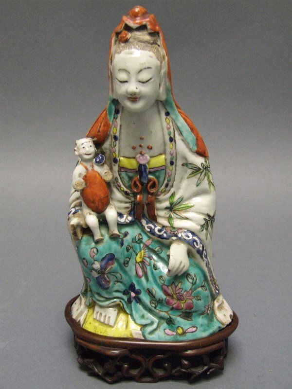 Chinese Porcelain Guan Yin Figure 18th C.: In a sitting position with a child on her knee decorated with colored enamels complete with a later carved wooden stand and British Antique Dealers decal to bottom. In good condition with losses to ha
