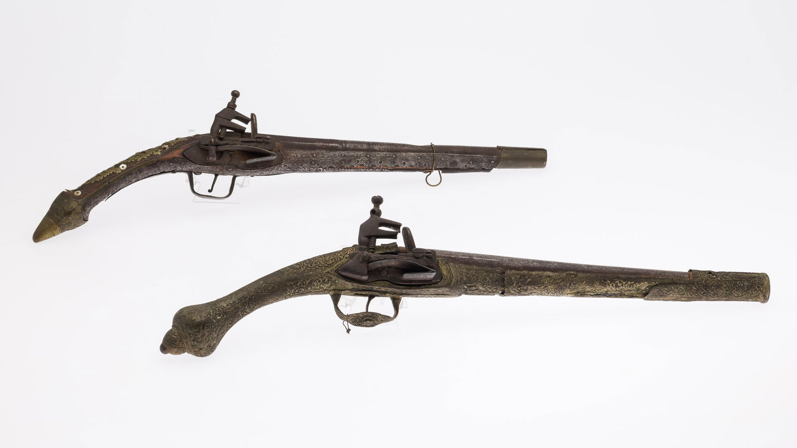 2 Middle Eastern Flint Lock Pistols: Property of a Diplomatic World-Traveling Family Description: 2 Middle Eastern Flint Lock Pistols, a 19 in. long pistol with pointed butt and scrolling design on applied metal surface (hole in top