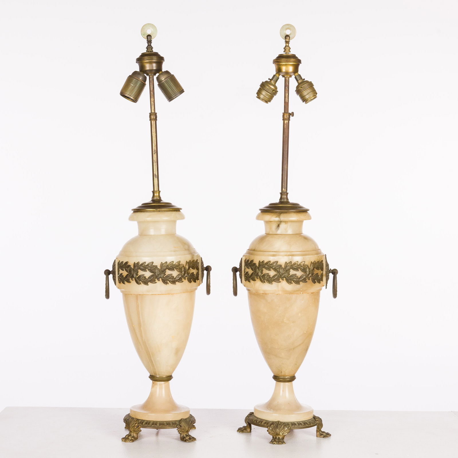 Pair of French Alabaster Urn-Form Lamps: Description: Pair of French Alabaster Urn-Form Lamps, the pair of alabaster urns mounted with metal handles and foliate accents on claw foot bases, mounted as lamps with European wiring.