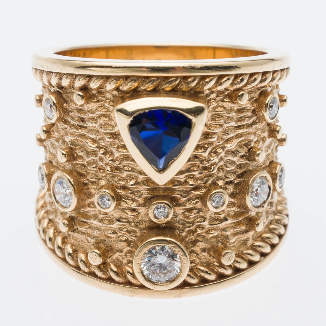 18K Gold, Sapphire and Diamond Ring, Size 8: Description: 18K Gold, Sapphire and Diamond Ring, Size 8, the wide textured yellow gold band featuring bezel set modified trillion cut sapphire and various round diamonds (approx. 0.61 cts), marked