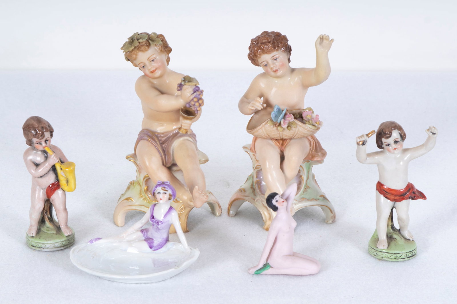 European Porcelain Figurines Putti & Girls (six): Four painted and glazed porcelain putti, two hallmarked underneath and two possibly hallmarked underneath, sizes from 5” tall to 3” tall; Together with a Bathing Beauty, painted and irides