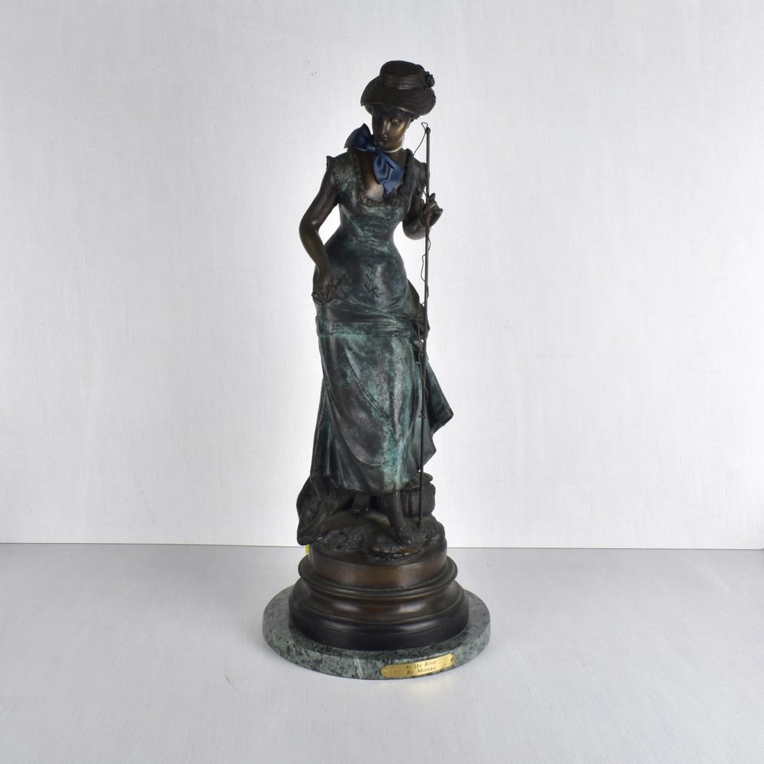 After Auguste Moreau, Fr. (1834-1917): After Auguste Moreau, French (1834-1917). Bronze figurine "At The River" cast by American Foundry. This sculpture depicts a Victorian woman holding a fishing pole in one hand and in the other hand hol
