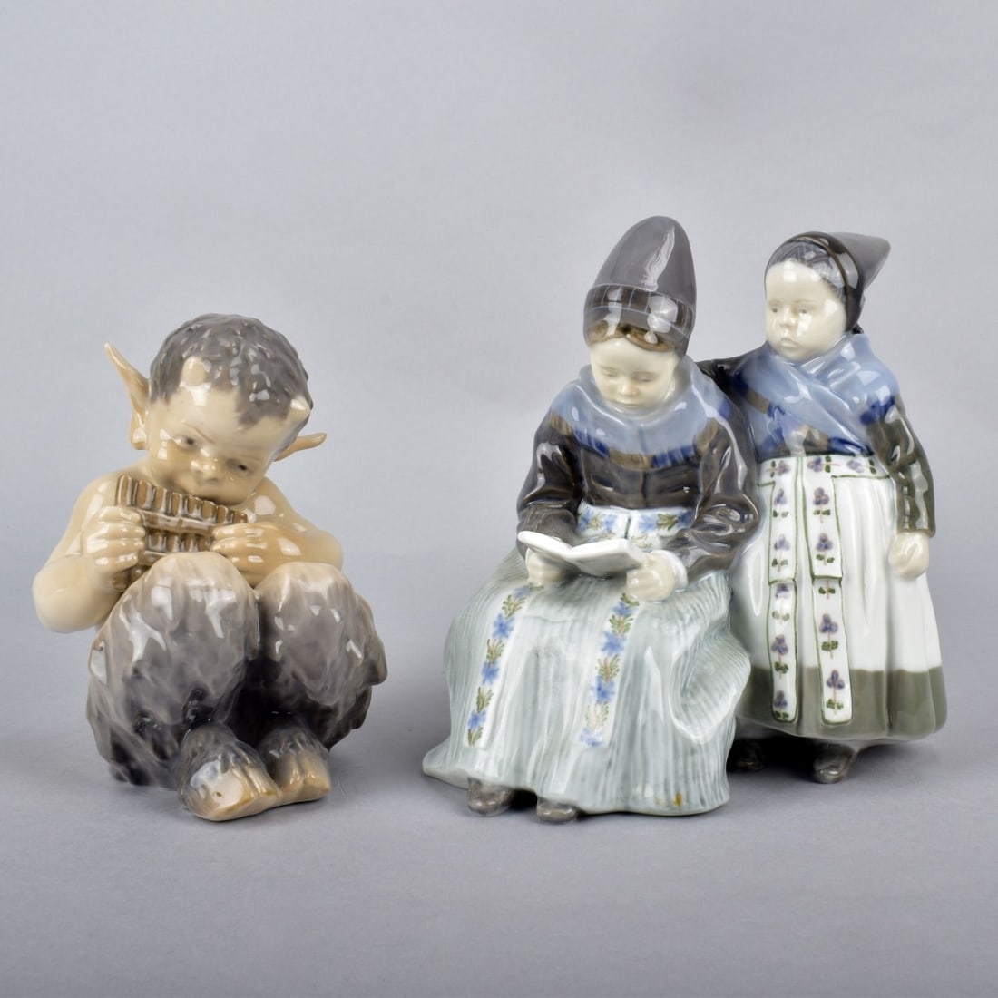 Two (2) Royal Copenhagen Porcelain Figurines: Two (2) Royal Copenhagen Porcelain Figurines. Dutch Amager Girls Reading #1395 measures 7"H x 6-1/2"W along with Fauna with Flute Figurine #1936 measures 5-3/4"H x 6"L. Kodner Galleries offers phone a
