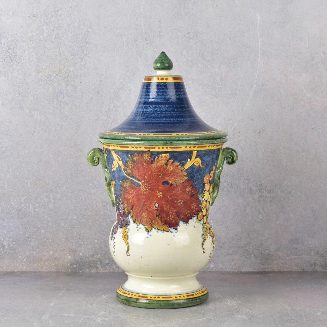 Vintage Leona Italian Porcelain Vase with Cover: Vintage Leona Italian Porcelain Vase with Cover. Decorated with grape leaves and grape clusters hand painted in bright colors with attached handles and an acorn finial. Signed "Leona" to the side and 