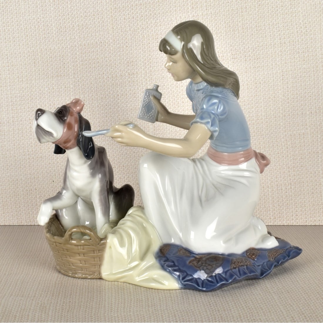 Lladro Figurine of a Girl and Dog: Lladro Porcelain Figurine of a Young Girl Attending to Her Sick Dog Sitting in a Basket. Properly marked on the bottom. Measures: 7-1/4" H8" W, 6" D. Kodner Galleries offers phone and absentee bidding