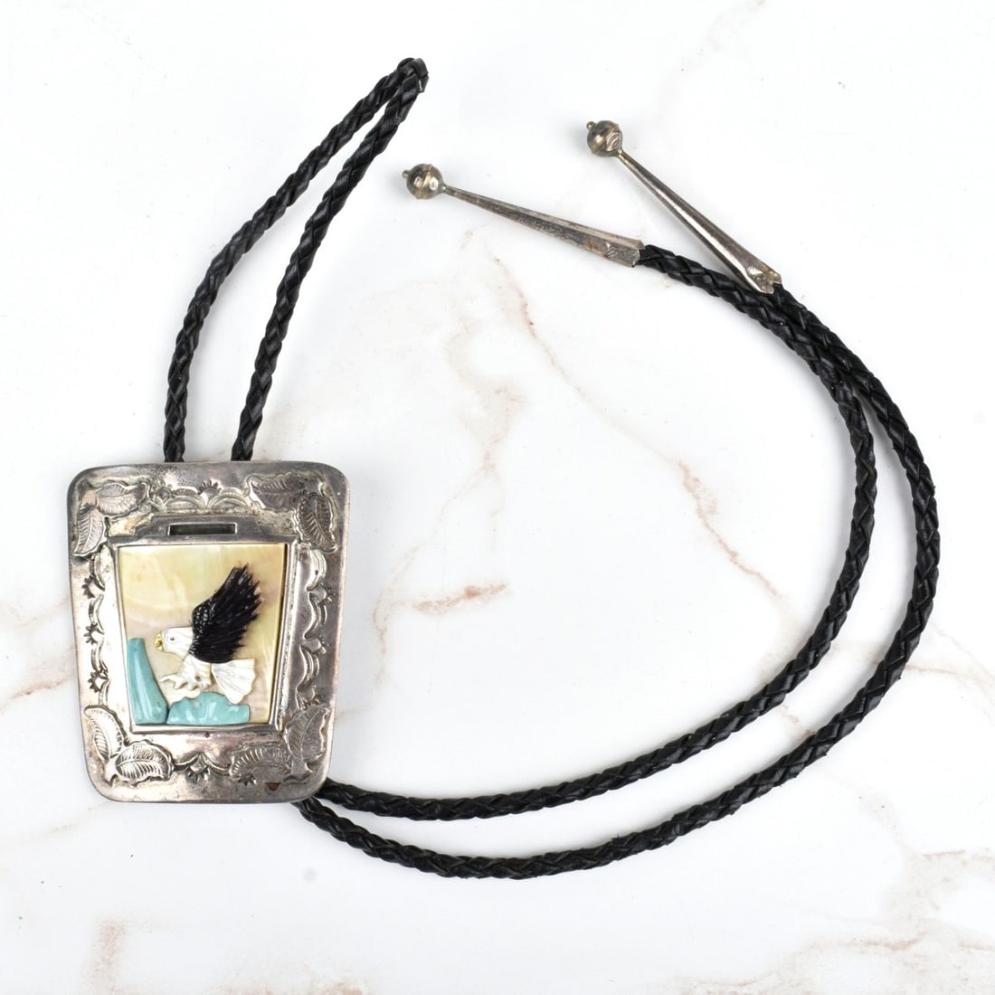 Zuni Hardstone and Silver Bolo Tie: Vintage Jones Quam Zuni Mother of Pearl, Black Onyx and Turquoise Inlaid Silver Bolo Tie with Braided Leather. Signed, stamped Zuni. Slider measures 2-3/4" H, 2-3/8" W; tie measures 38" L. Approx. gro