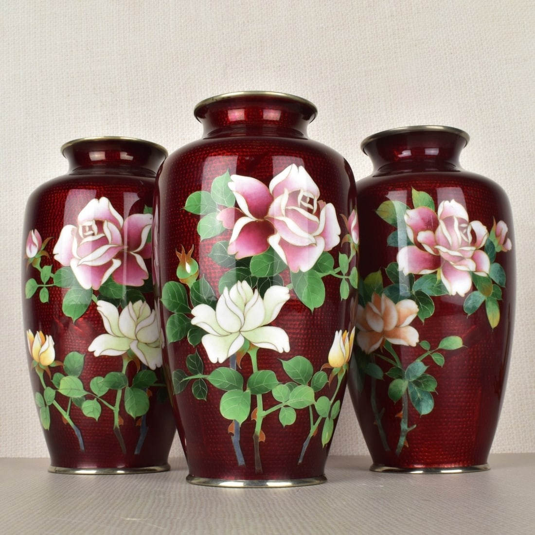 Japanese Cloisonne Vases: Three (3) Japanese Cloisonne Pigeon Blood Red Ginbari Foil Vases Decorated with Large Roses. Mid 20th century. Measures: 7-1/16" H, 4" D. Kodner Galleries offers phone and absentee bidding at a low, f