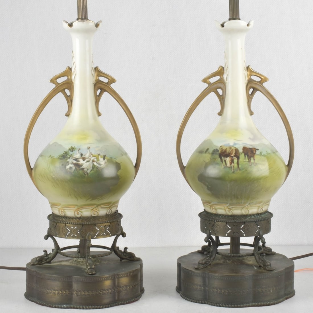 Pair of Porcelain Urn Shape Lamps: Pair of Porcelain Urn Shape Table Lamps on Brass Bases. Circa 1940's. Decorated with rural pastoral scenes of grazing cows and geese. Porcelain body of lamps measure: 12-1/4" high, 7-1/2" wide, 6" dee