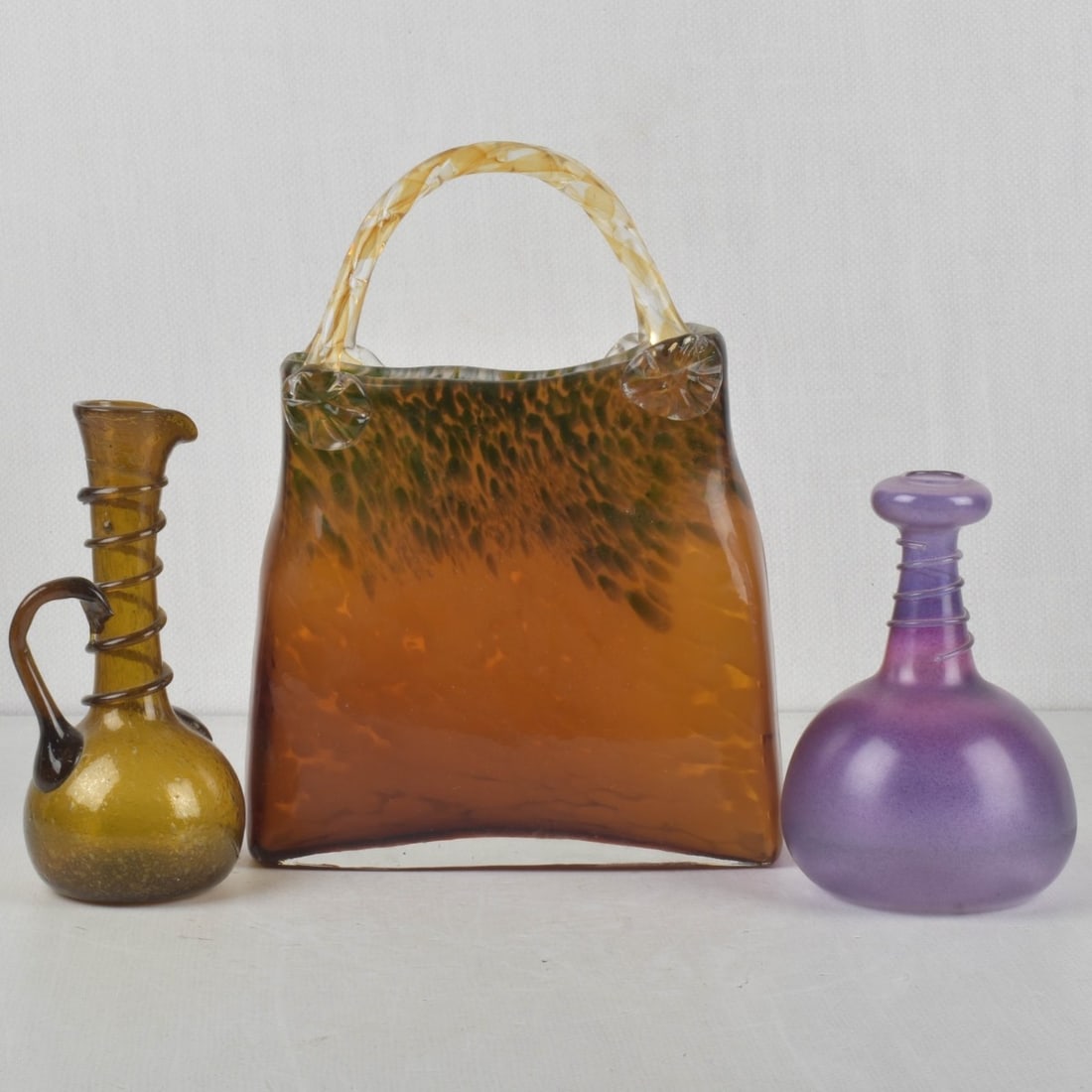 Modern Art Glass Pieces: Three (3) Modern Art Glass Pieces. Consisting of one (1) Murano glass purse in orange-black glass with attached handles, measures: 12" high, 8-1/2" wide, 2-3/4" deep, one (1) amber glass pitcher with 