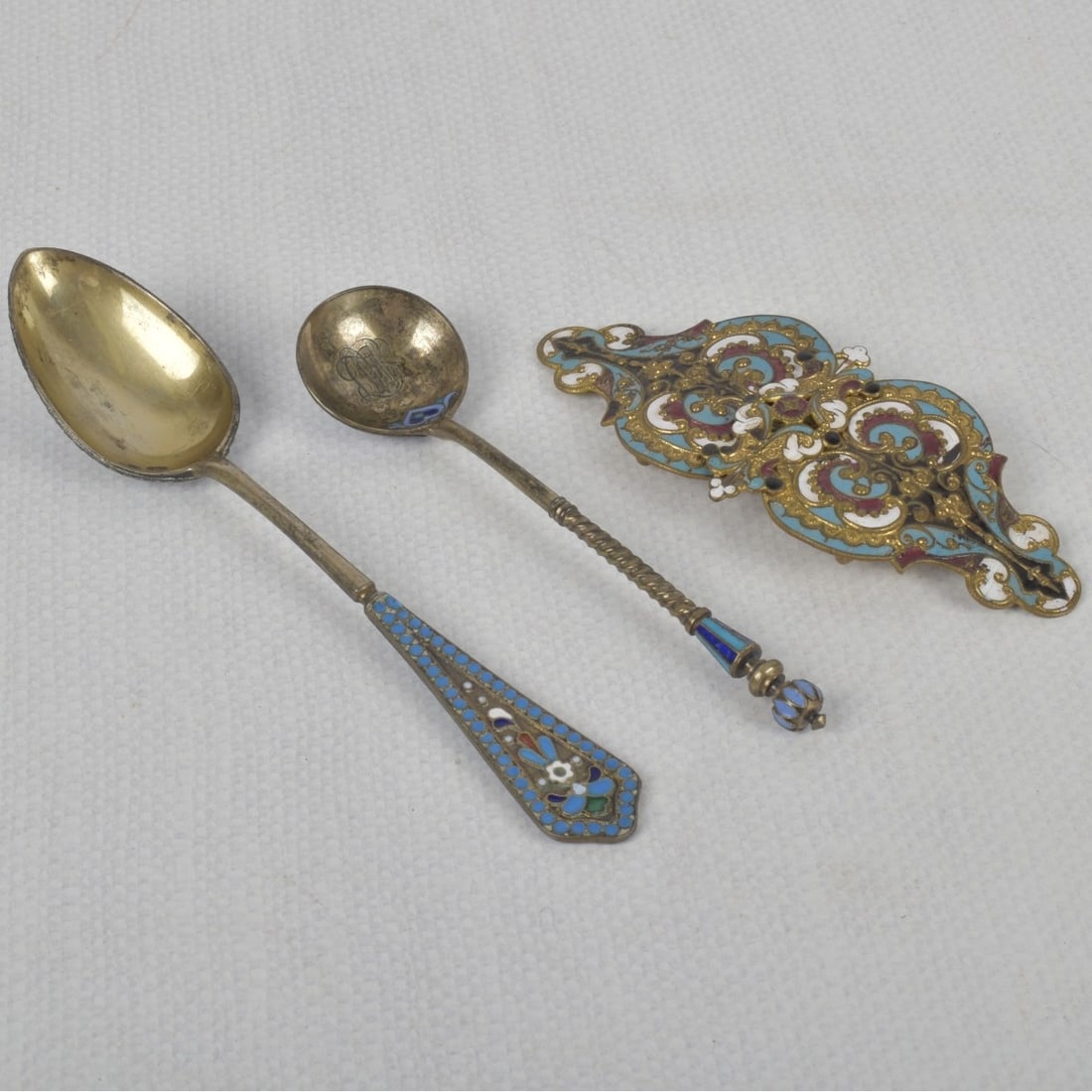 Russian Silver and Champleve: Russia Silver and Champleve Pieces. Consisting of one (1) spoon measures: 6-3/4" long, one (1) monogrammed spoon, measures: 5-1/4" long, one (1) belt buckle, measures: 4-3/4" long, 2" high, to