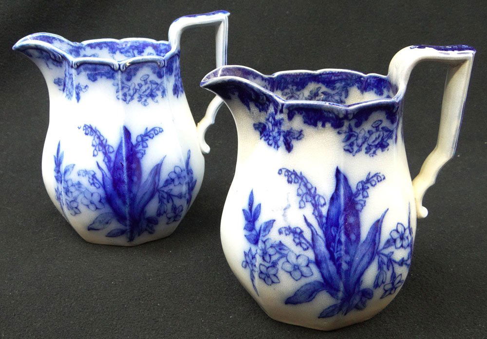 Two (2) Flow Blue Milk/Crème Pitchers in the Lily of: Two (2) Flow Blue Milk/Crème Pitchers in the Lily of the Valley Pattern. Unsigned. Normal Handling Ware, Minor Roughness on the Base of One Pitcher Otherwise in Good Condition. Shipping $42.00
