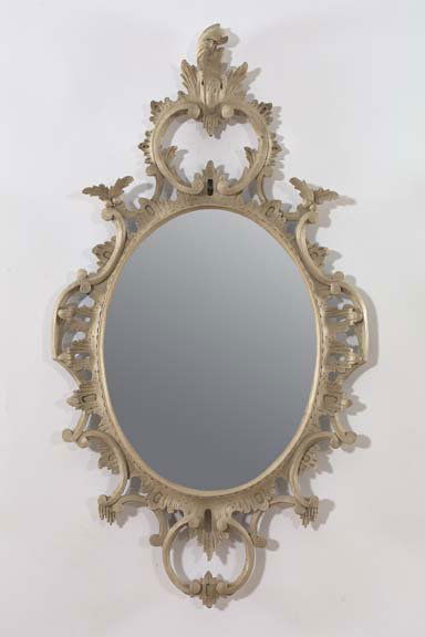 English Carved Wood Mirrors: Pair of English Carved and Antique-Gray-Painted Wood Oval Looking Glasses, in the rococo style of Thomas Johnson (1714-1778), the frames elaborately pierced and carved, the tall crestings modeled as p