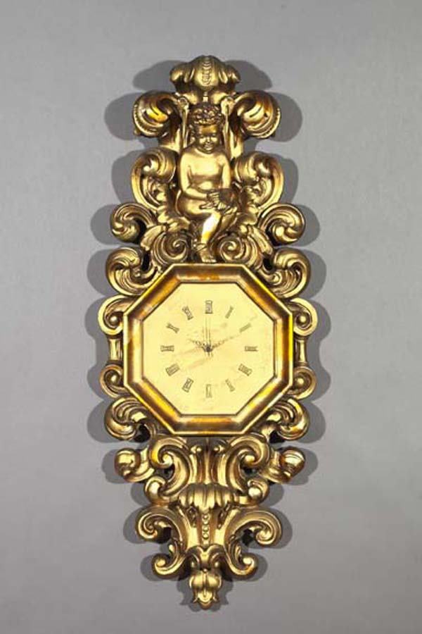 CARVED GILTWOOD WALL CLOCK: Large and Dramatic Carved Giltwood and Plaster Wall Clock, in the Roman Baroque style, the face octagonal, with relief Roman chapters, surmounted by a putto perched amidst boldly modeled foliate scrol