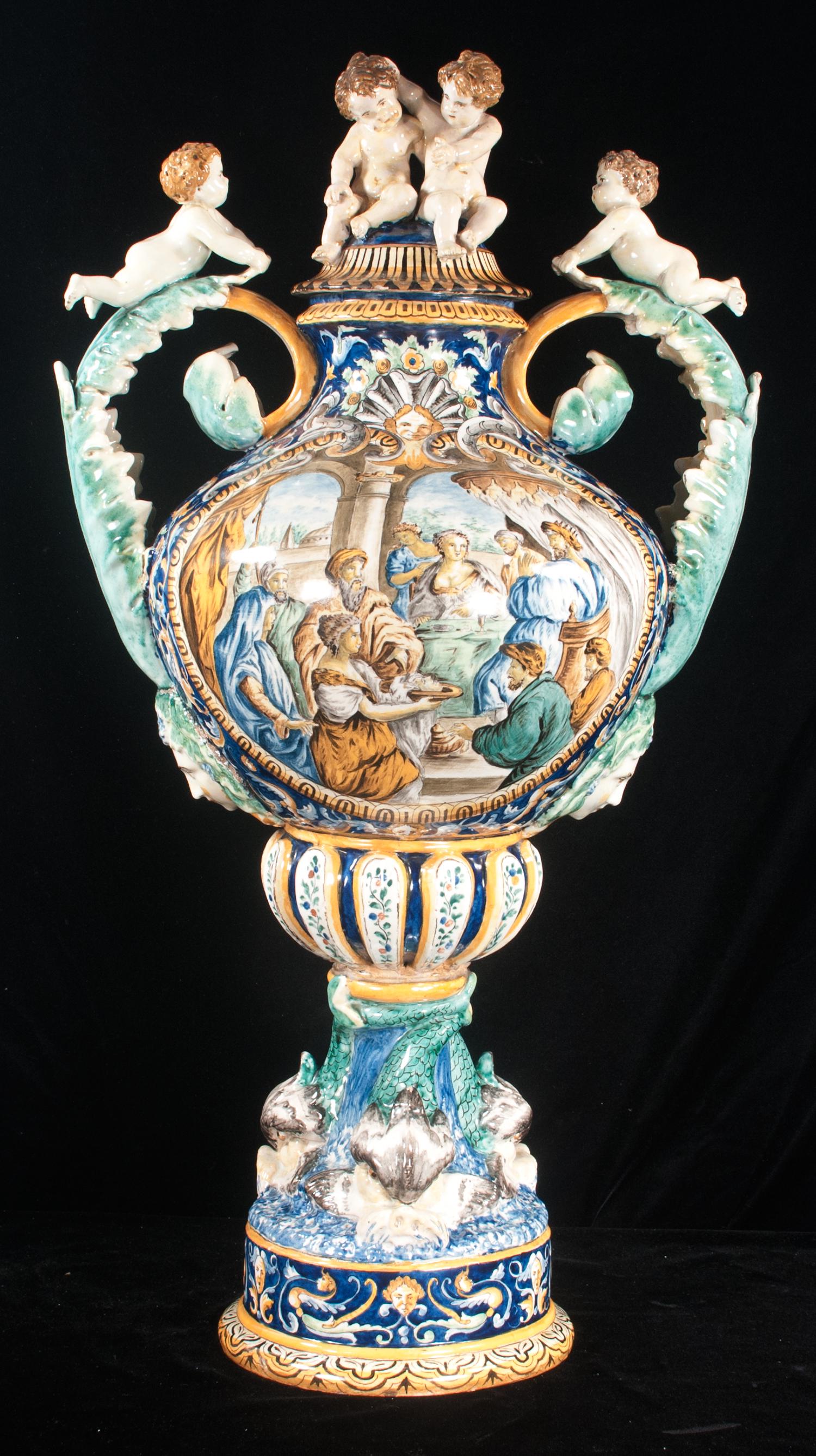 Palace size Italian Majolica capped urn: Palace size Italian Majolica capped urn, the lid has two putti mounted on top and each leaf formed handle has a putti on top, body has panel decoration with a group of figures and supported by three d