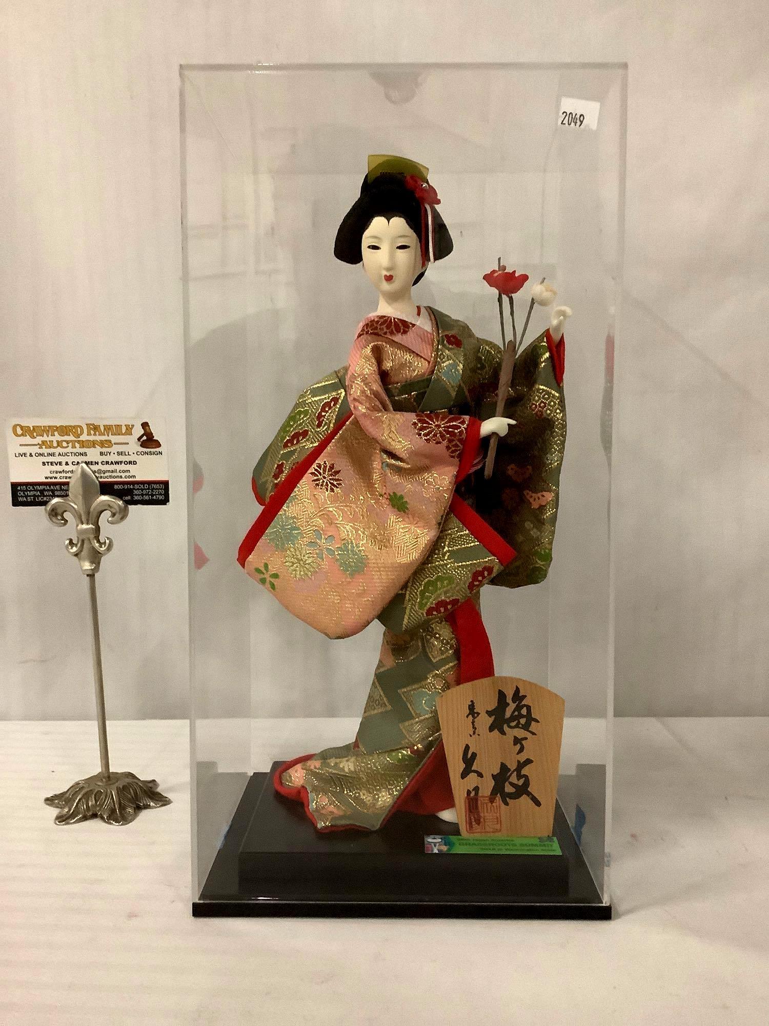 2018 Japanese female doll holding a plum blossom in: 2018 Japanese doll holding a plum blossom in glass display box, possibly Princess Asagao from the Tale of Genji, from the Japan-America Society, approx. 7.5x6.5x14.5 inches.