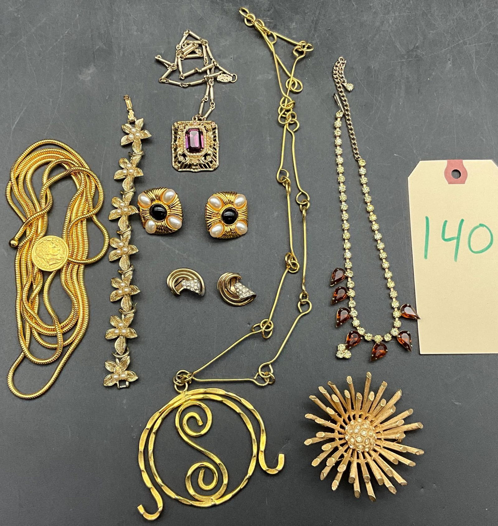 Gold-Toned Costume Jewelry Including Coro and Napier: Gold-toned costume jewelry consisting of necklaces, brooches, earrings, and bracelets. Notable pieces include a brooch marked 'Coro,' a pair of earrings stamped 'Napier,' a large initi