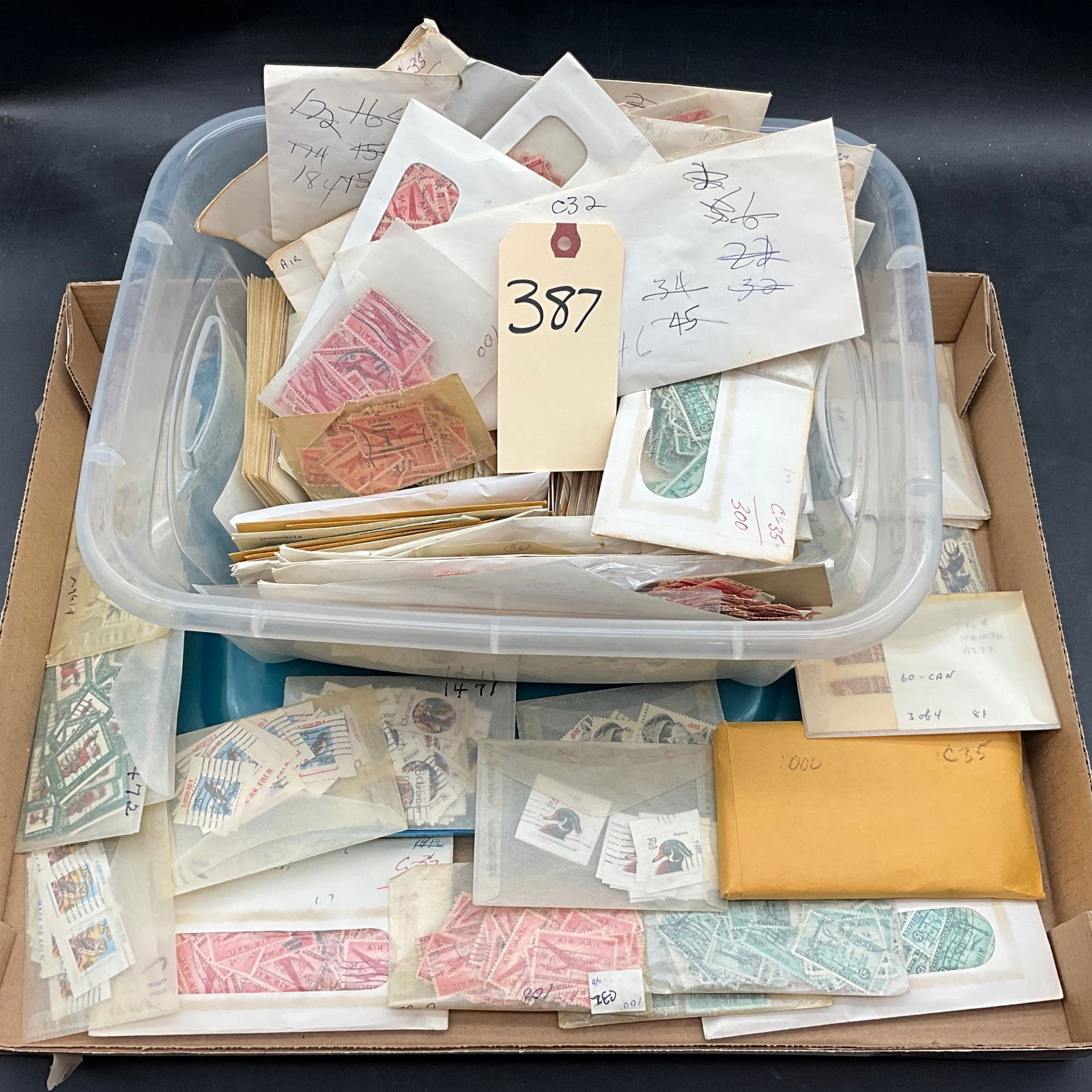 An Assortment of Vintage United States Postage Stamps: This collection features an assortment of vintage United States postage stamps organized within glassine and paper envelopes, a plastic bin, and a cardboard tray. The lot includes various