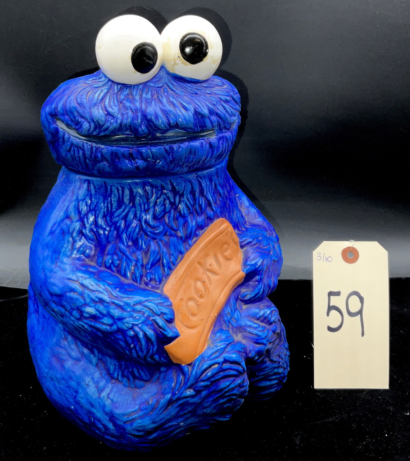 Vintage Sesame Street Cookie Monster Ceramic Cookie Jar, c. 1970s: This vintage Sesame Street molded ceramic cookie jar depicts the character Cookie Monster in a seated pose holding a brown biscuit embossed with the word "Cookie." The two-piece vessel features a text