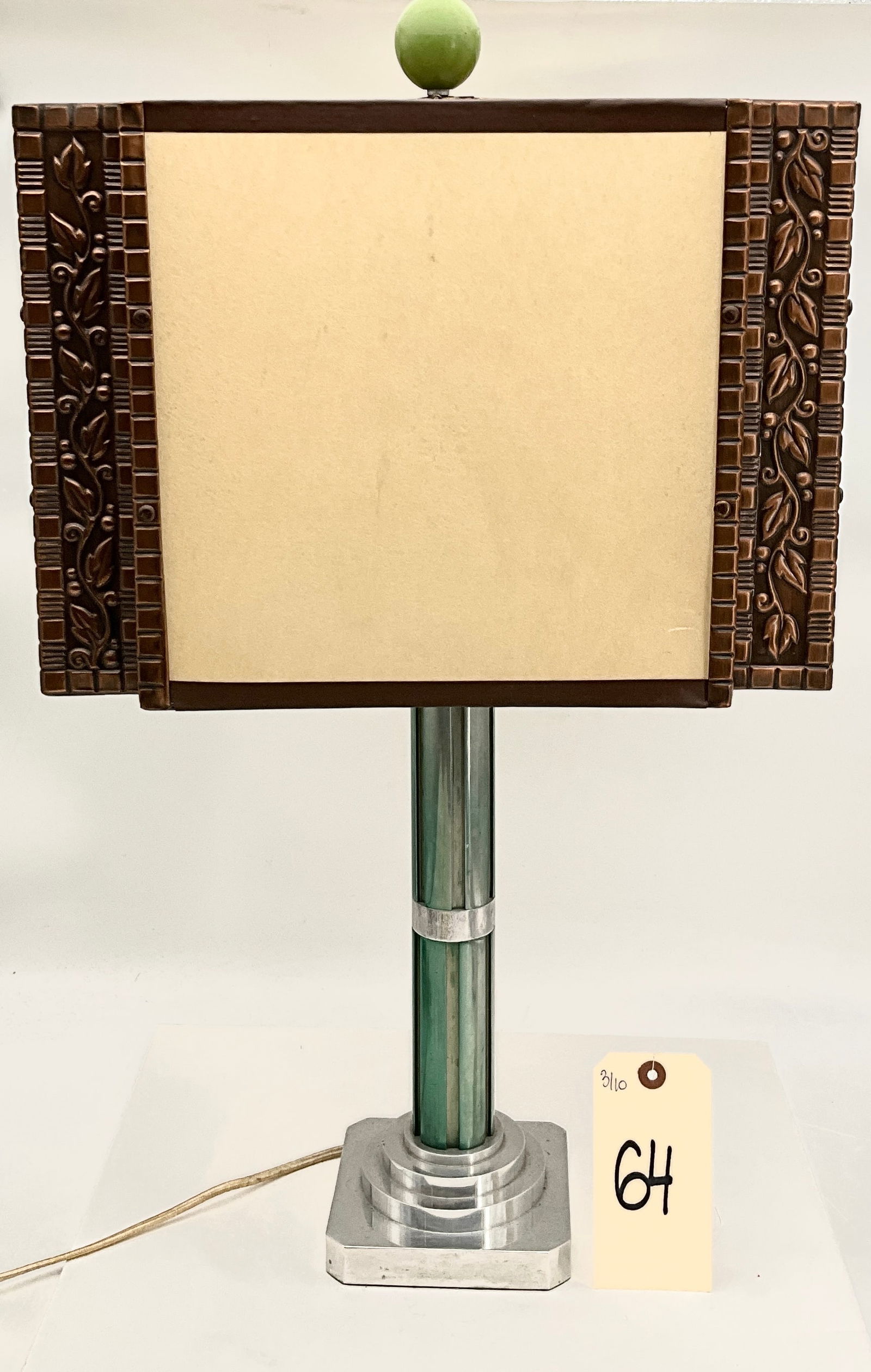 American Machine Age Art Deco Table Lamp: This Art Deco table lamp features a geometric, stepped skyscraper base in polished metal accented by a central column of vertical green glass rods. The rectangular shade consists of a patinated metal
