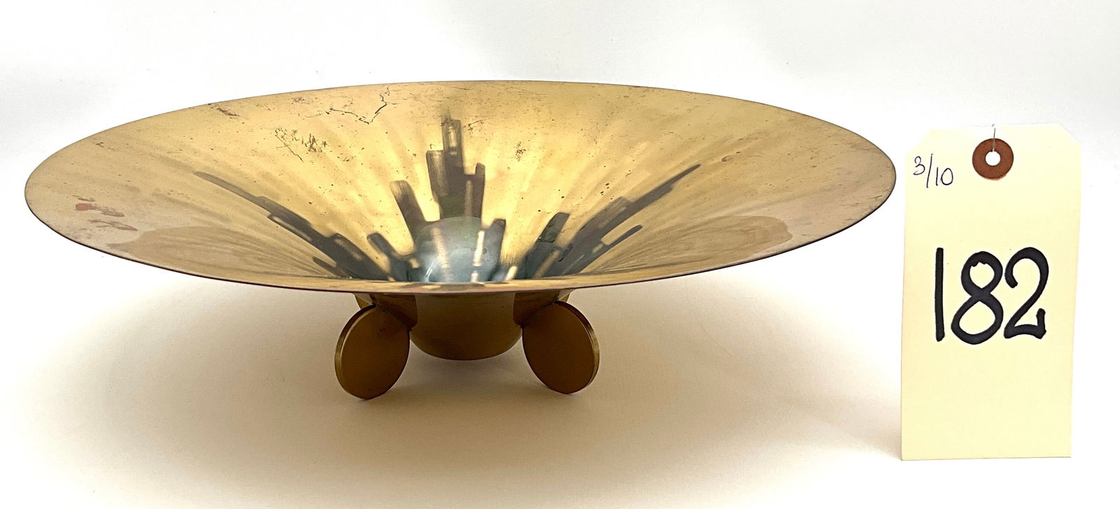 A Mid-Century Modern WMF Ikora Brass Centerpiece Bowl: This mid-century modern WMF Ikora brass centerpiece bowl features a shallow, wide-rimmed conical body elevated by four circular disc feet. The interior is decorated with a central, black abstract geom