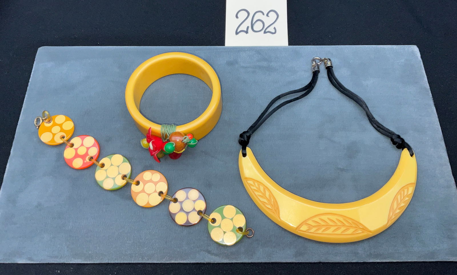 Three Vintage Bakelite Jewelry Pieces, Polka Dot and Carved Leaf Motifs: This three-piece collection of vintage Bakelite jewelry features a carved crescent necklace, a multi-colored polka dot link bracelet, and a charm-adorned bangle. The necklace showcases a yellow