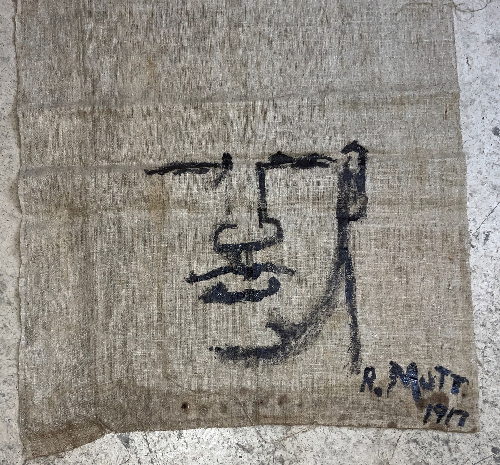 R. Mutt, Painted Textile Fragment, Minimalist Face Motif, 1917: This minimalist work features a stylized human face rendered in thick dark pigment on a coarse, light-brown woven textile. The piece is prominently signed R. MUTT and dated 1917 in the lower right