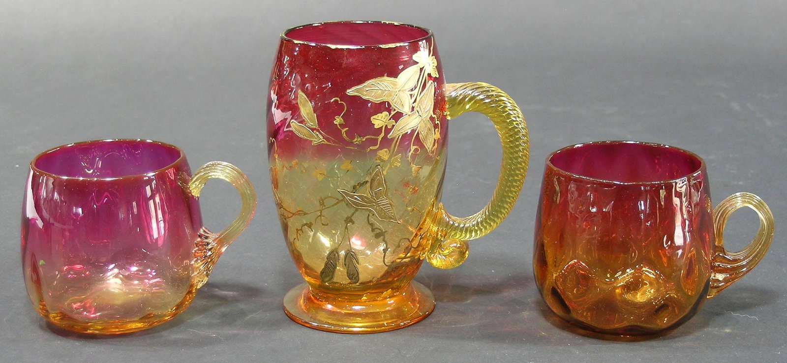 THREE AMBERINA GLASS DRINKING VESSELS: comprising a swirl optic mug having gilt floral, leaf and vine decoration, with twist handle, 4 1/2" high, and two punch cups, both quilted and having reeded handles, 2 1/2" high