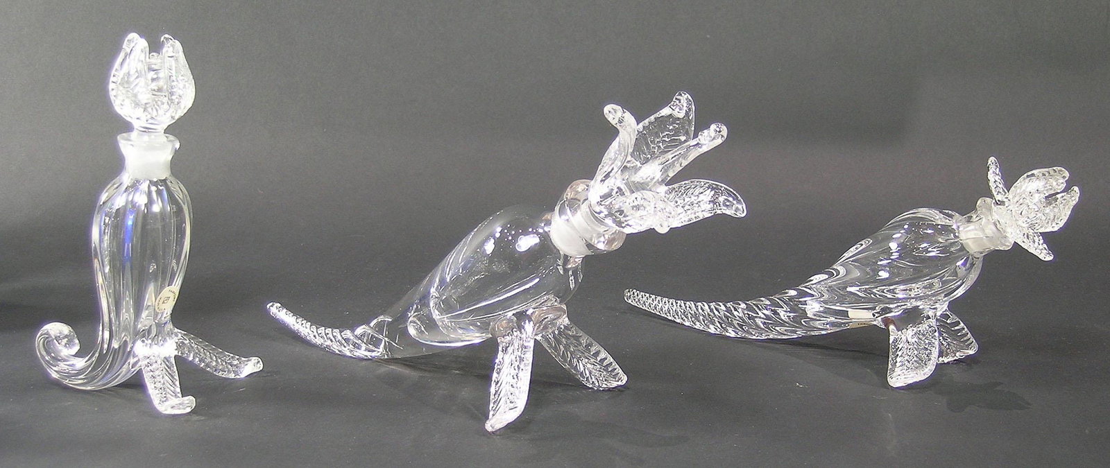 THREE PAIRPOINT GLASS CORNUCOPIA PERFUME BOTTLES: colorless glass, all having flower form stoppers and raised on leaf form legs, two being horizontal with one having twisting air bubbles in the base of the cornucopia, the longest being 10 1/2" long, 