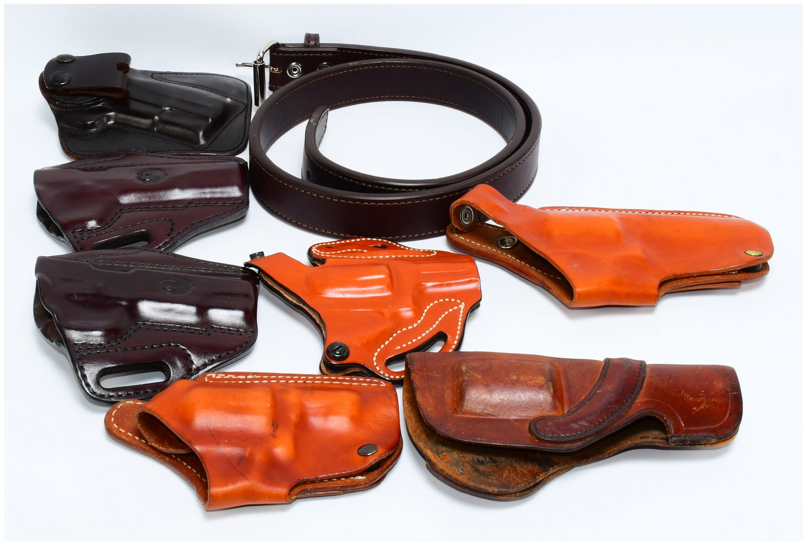 A Collection of Leather Firearm Holsters and a Belt: A collection of seven molded leather firearm holsters and one leather belt. The holsters are crafted from various shades of brown leather, ranging from dark burgundy/cordovan to lighter saddle-tan, wi
