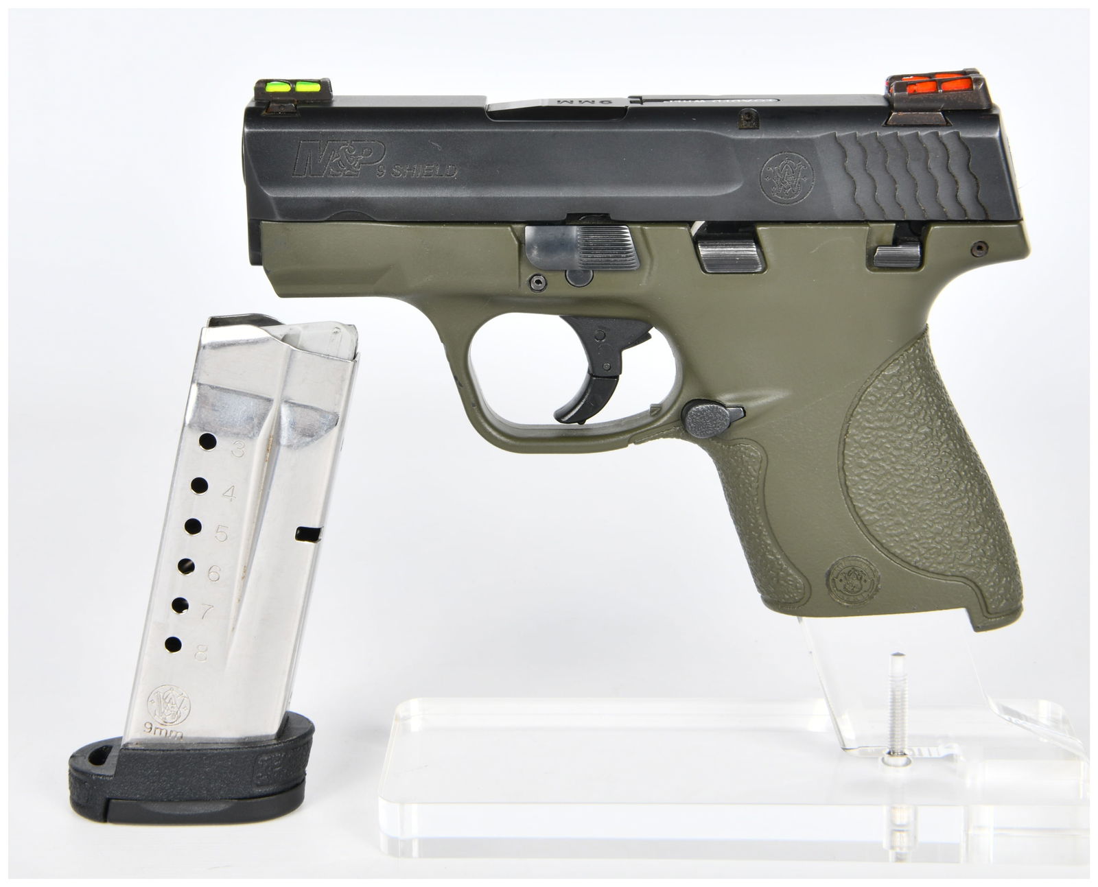 A Smith & Wesson M&P 9 SHIELD 9mm Semi-Automatic Pistol with OD Green Frame and Fiber Optic Sights: A modern, compact, semi-automatic pistol, identified through web intelligence as a Smith & Wesson M&P 9 SHIELD. The pistol features an olive drab green polymer frame and a black metal slide. It is equ
