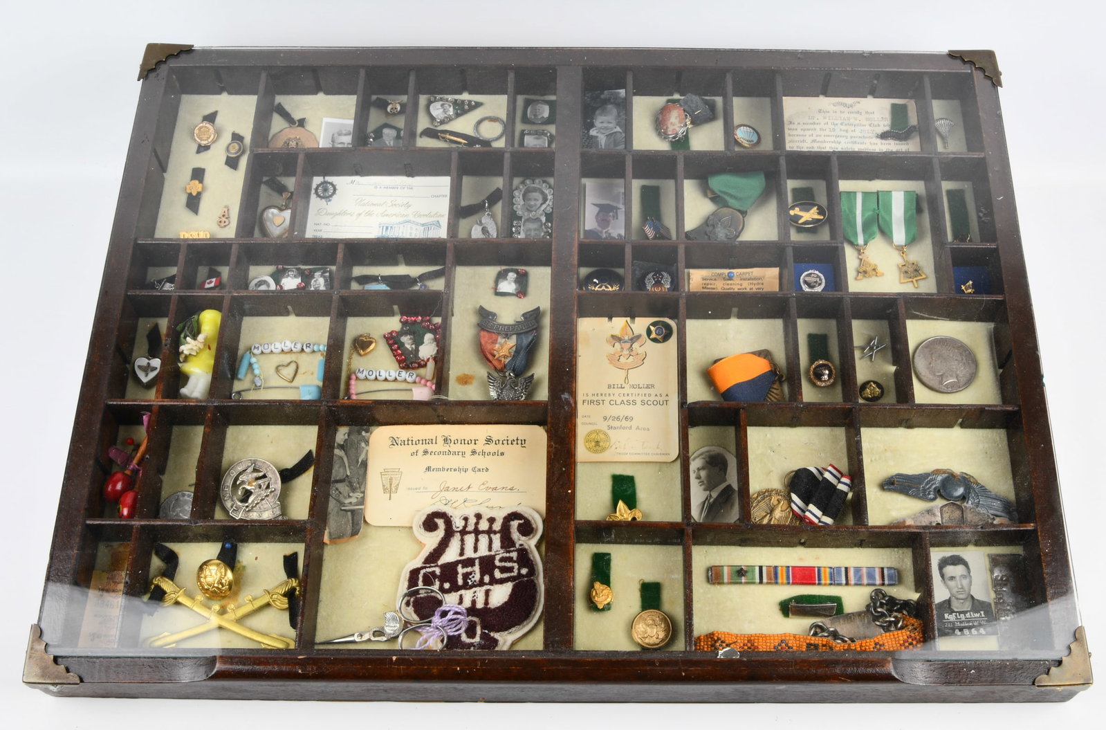 Moller Family Collection of Military, Boy Scout, and Personal Memorabilia in Display Case: A framed wooden display case, likely a repurposed printer's type tray, with a glass top and metal corner protectors, filled with a diverse collection of personal memorabilia. The contents, arranged in