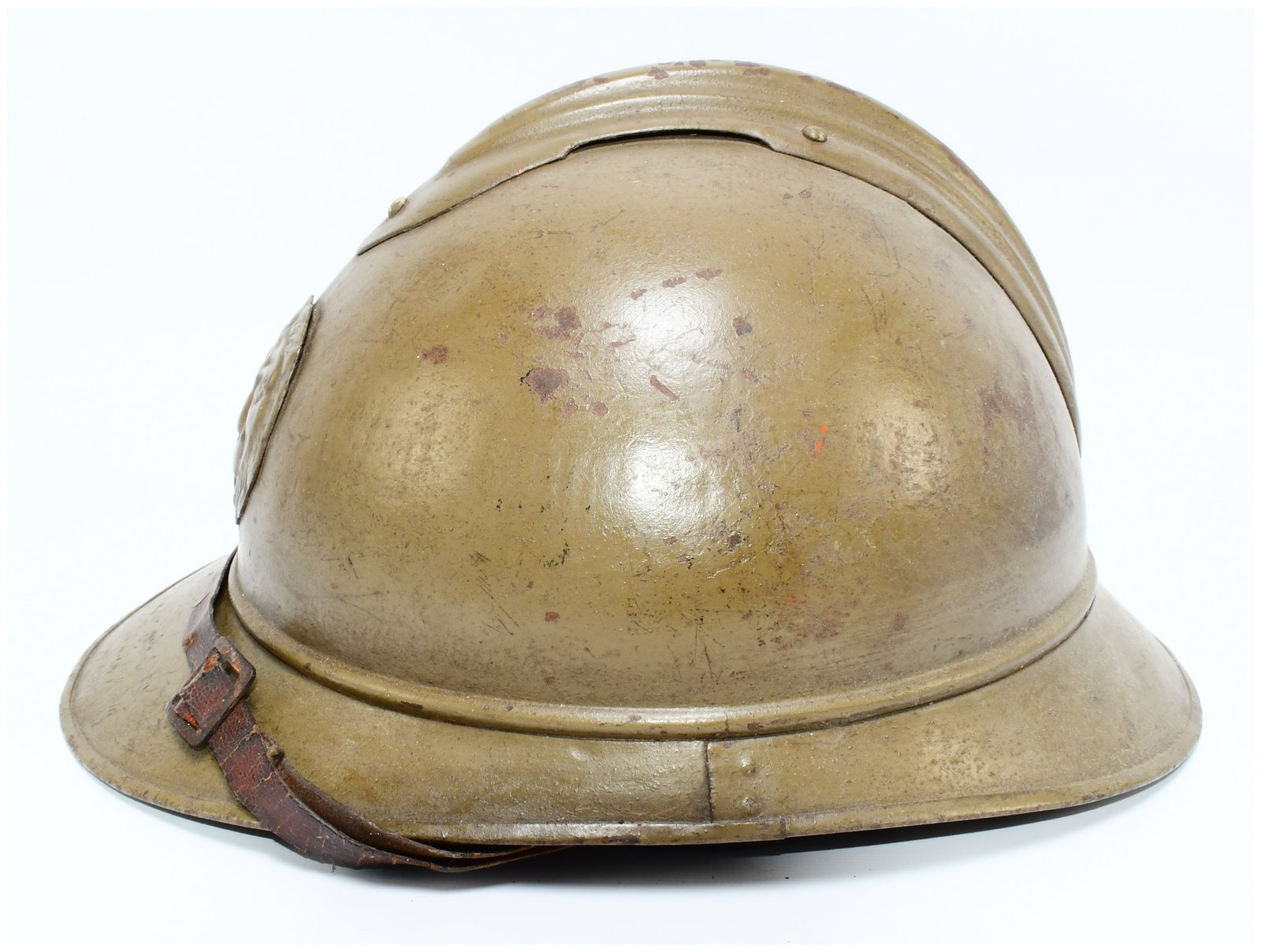 Belgian WWI M1915 Adrian Helmet: A Belgian WWI M1915 Adrian Helmet, constructed from olive drab-painted metal with a distinctive flaring brim and a raised central crest. An applied metal emblem depicting a lion's head in relief is af