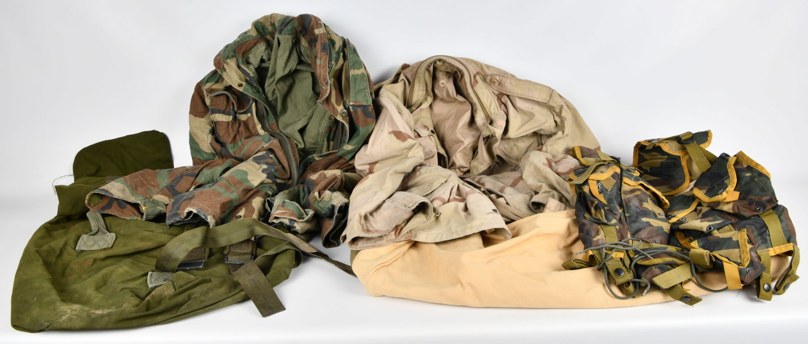 A Collection of USGI M65 Woodland and Desert Camouflage Field Gear: A collection of military-style field gear, comprising two M-65 type field jackets, a tactical vest, and two canvas duffel bags. The first jacket is a USGI M65 Woodland Camouflage Field Jacket, labeled