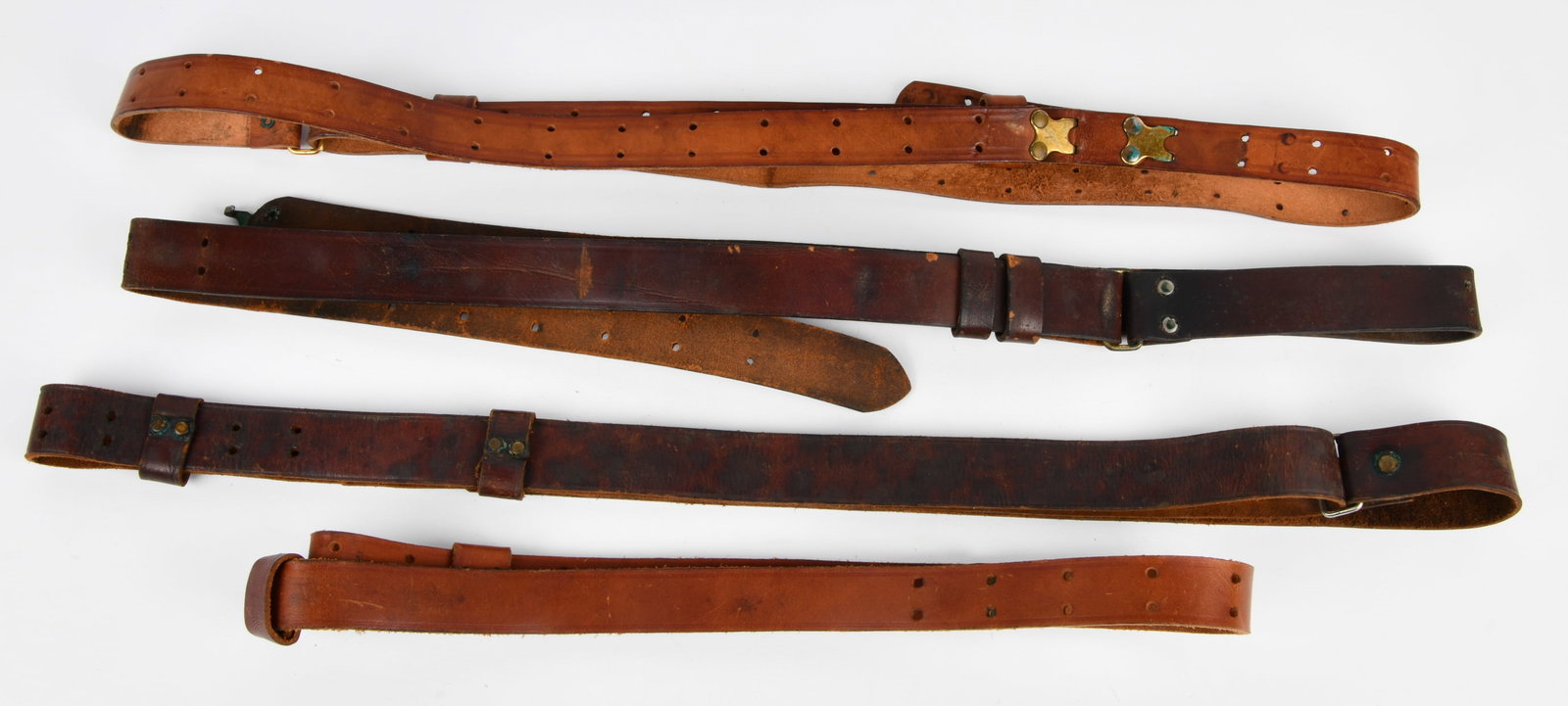 A Group of Four Vintage Leather Military Rifle Slings: A group of four vintage leather rifle slings, characteristic of military or outdoor equipment. The straps vary in color from light tan to dark reddish-brown. Each strap features a series of punched ho