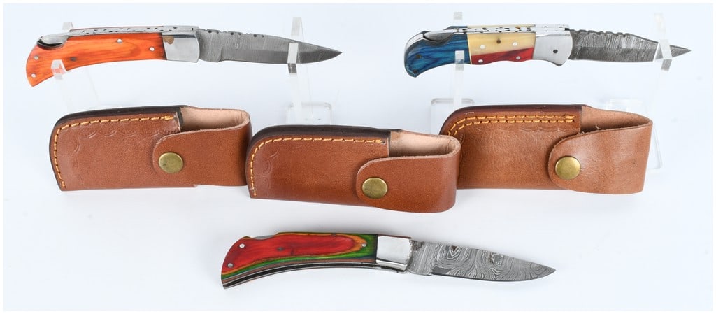 A Trio of Modern Handmade Damascus Steel Folding Pocket Knives with Sheaths: A collection of three modern handmade folding pocket knives, each featuring a blade constructed of Damascus steel, distinguished by visible wavy patterns resulting from the forging process. The knives