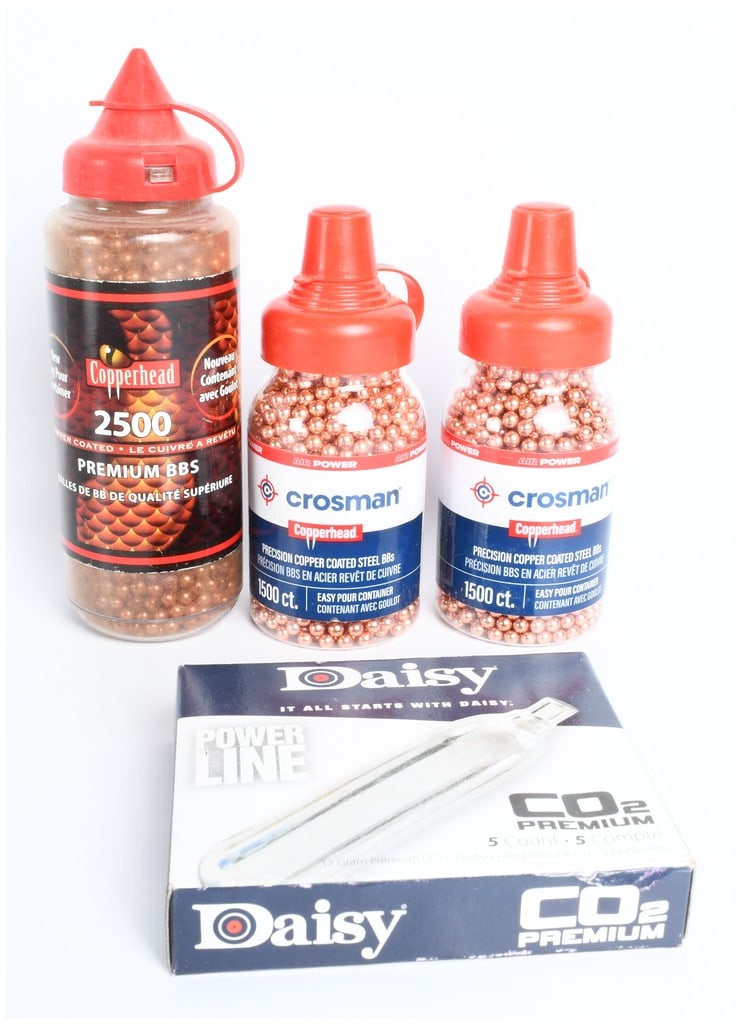 A Group of Crosman Copperhead BBs and Daisy Power Line CO2 Cylinders: A group of airgun accessories comprising Crosman Copperhead copper-coated steel BBs and Daisy Power Line CO2 cylinders. The collection includes a large plastic dispenser of Copperhead Premium BBs