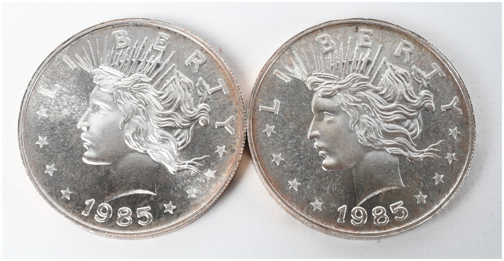 A Pair of 1985 Peace Dollar Design 1 Troy Ounce .999 Fine Silver Trade Units: This lot consists of two struck silver bullion rounds featuring an obverse design styled after the United States Peace Dollar. The obverse of each round depicts a profile of Liberty facing left, with 