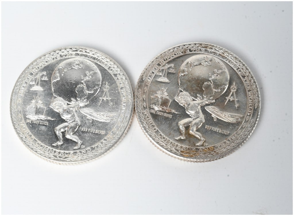 A Pair of .999 Fine Silver 1 Troy Ounce World Commerce Bullion Rounds: This lot consists of two silver bullion rounds featuring the World Commerce and Trade design. The obverse of each round depicts a muscular figure holding a globe, surrounded by the inscriptions SILVER