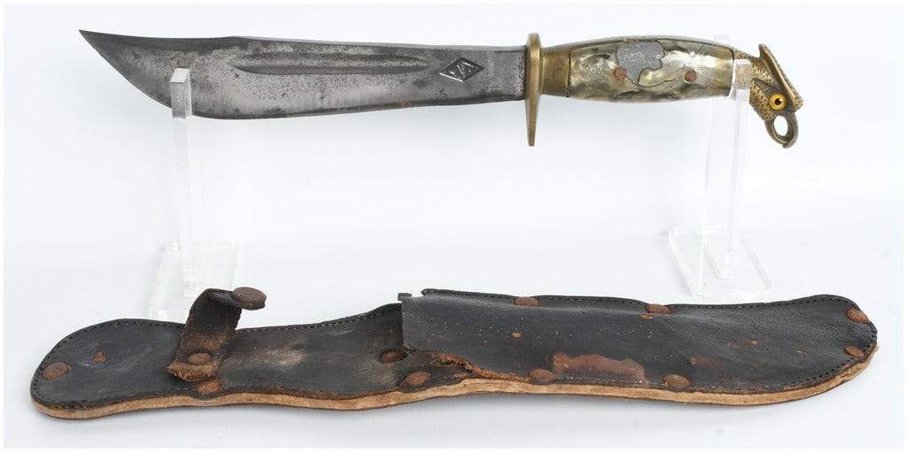 Mexican Eagle-Head Fighting Knife with Leather Scabbard: A Mexican fighting knife featuring a large steel clip-point blade with a single fuller running along the upper third of each side. The blade is stamped on the ricasso with a diamond-shaped mark contai