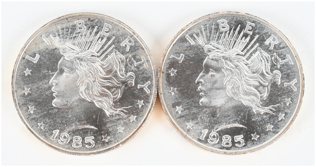 A Pair of 1985 Peace Dollar Design 1 Troy Ounce .999 Fine Silver Trade Units: This lot features two silver bullion rounds, each containing 1 troy ounce of .999 fine silver. The design on the obverse is inspired by the United States Peace Dollar, showing a profile of Liberty wit