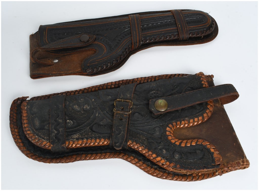 A Pair of Tooled Leather Western Style Revolver Holsters: This lot features a pair of vintage leather western-style revolver holsters showcasing distinct styles of traditional leathercraft. The first holster is crafted from dark brown leather with tooled lin