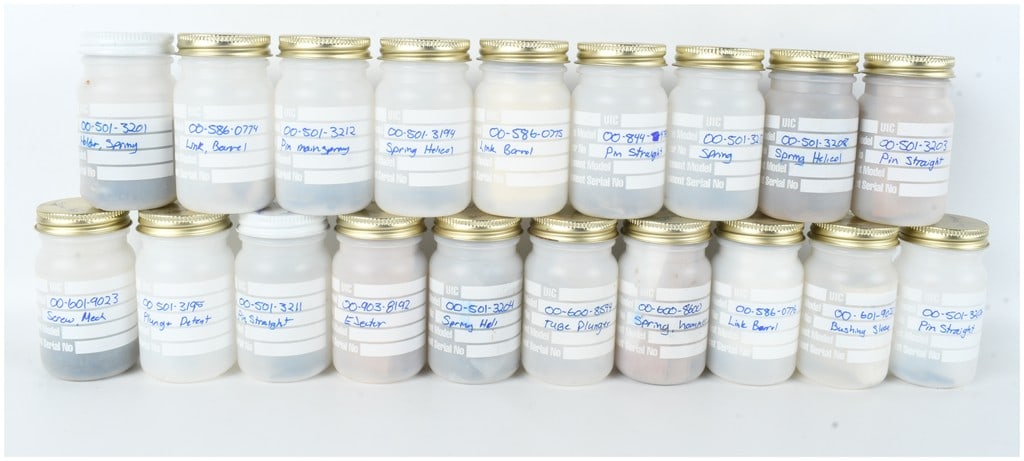 A Collection of 19 Jars of Assorted Firearm Components and Replacement Parts: This lot features 19 white plastic storage jars containing a variety of small firearm components and replacement parts. Each jar is topped with a screw-cap lid, most of which are gold-toned metal. The