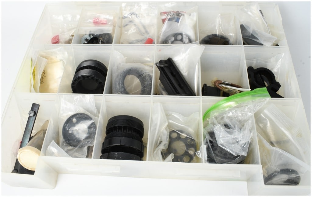 A Collection of AR-15 Rifle Parts and Armorer Components: A collection of AR parts and armorer components housed in a multi-compartment translucent plastic storage case. This assortment includes various black-finished metal mounting hardware such as Picatinn