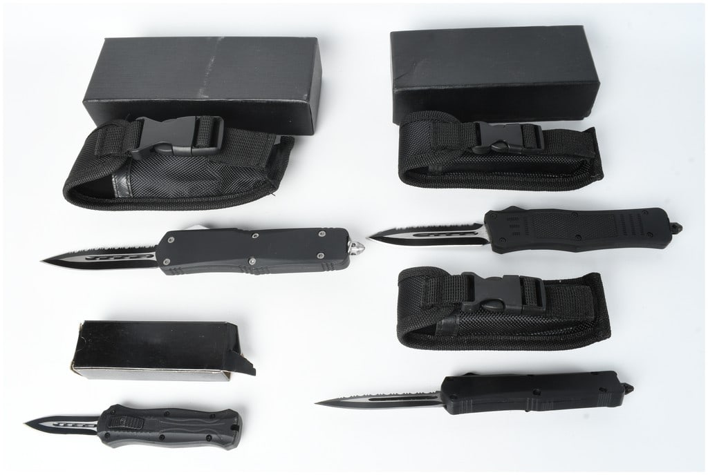 A Collection of Four Gunner Gear Out-the-Front Automatic Knives: This lot consists of a collection of four Gunner Gear out-the-front automatic knives accompanied by three black nylon belt sheaths with plastic quick-release buckles and three black storage boxes. The