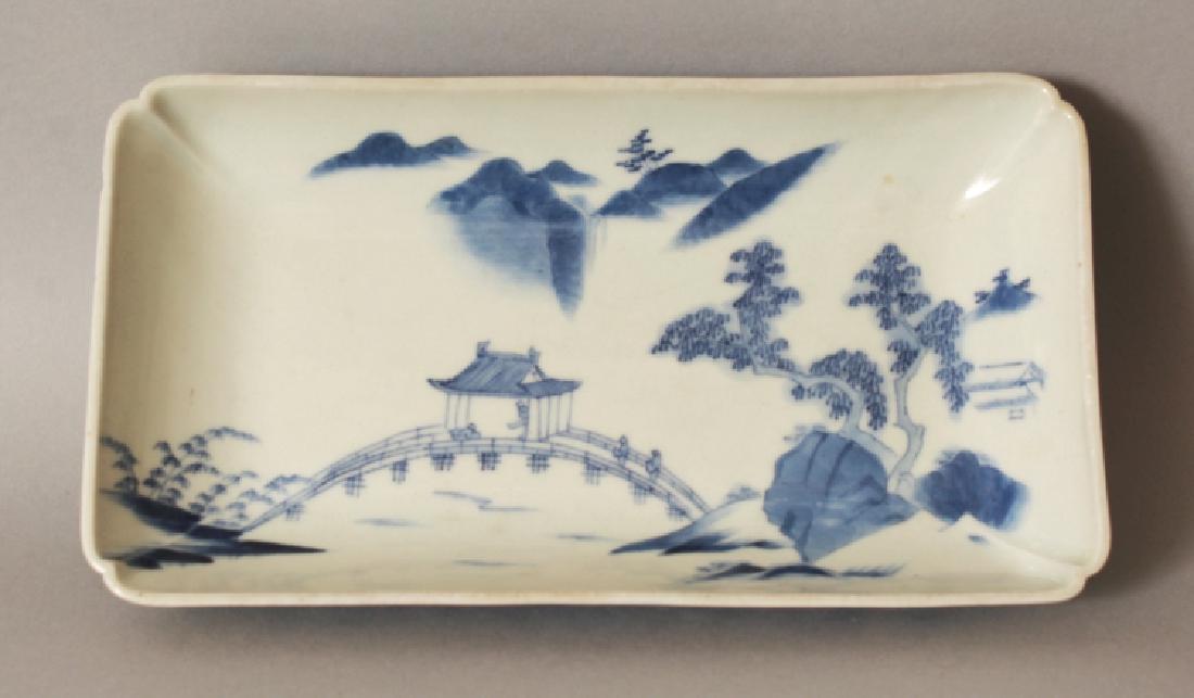 A GOOD QUALITY 18TH/19TH CENTURY NABESHIMA BLUE & WHITE: A GOOD QUALITY 18TH/19TH CENTURY NABESHIMA BLUE & WHITE RECTANGULAR PORCELAIN DISH, with indented corners and comb foot, the interior painted with a river bridge in a mountainous landscape setting, 8.