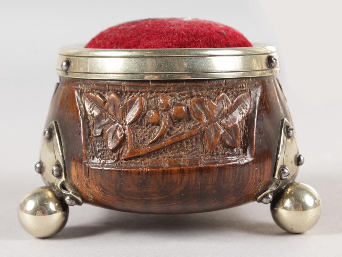 AN IRISH CARVED WOOD CIRCULAR PIN CUSHION, carved with: AN IRISH CARVED WOOD CIRCULAR PIN CUSHION, carved with fruiting vines, with plated rim and three ball feet. 2.5ins diameter.