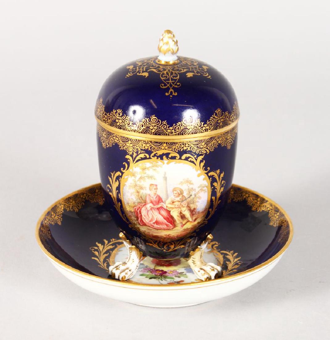 A 19TH CENTURY MEISSEN BLUE GROUND CUP, COVER AND: A 19TH CENTURY MEISSEN BLUE GROUND CUP, COVER AND STAND, the cup edged in gilt and painted with an oval, a young lady and boy playing a mandolin. Cross swords mark in blue.