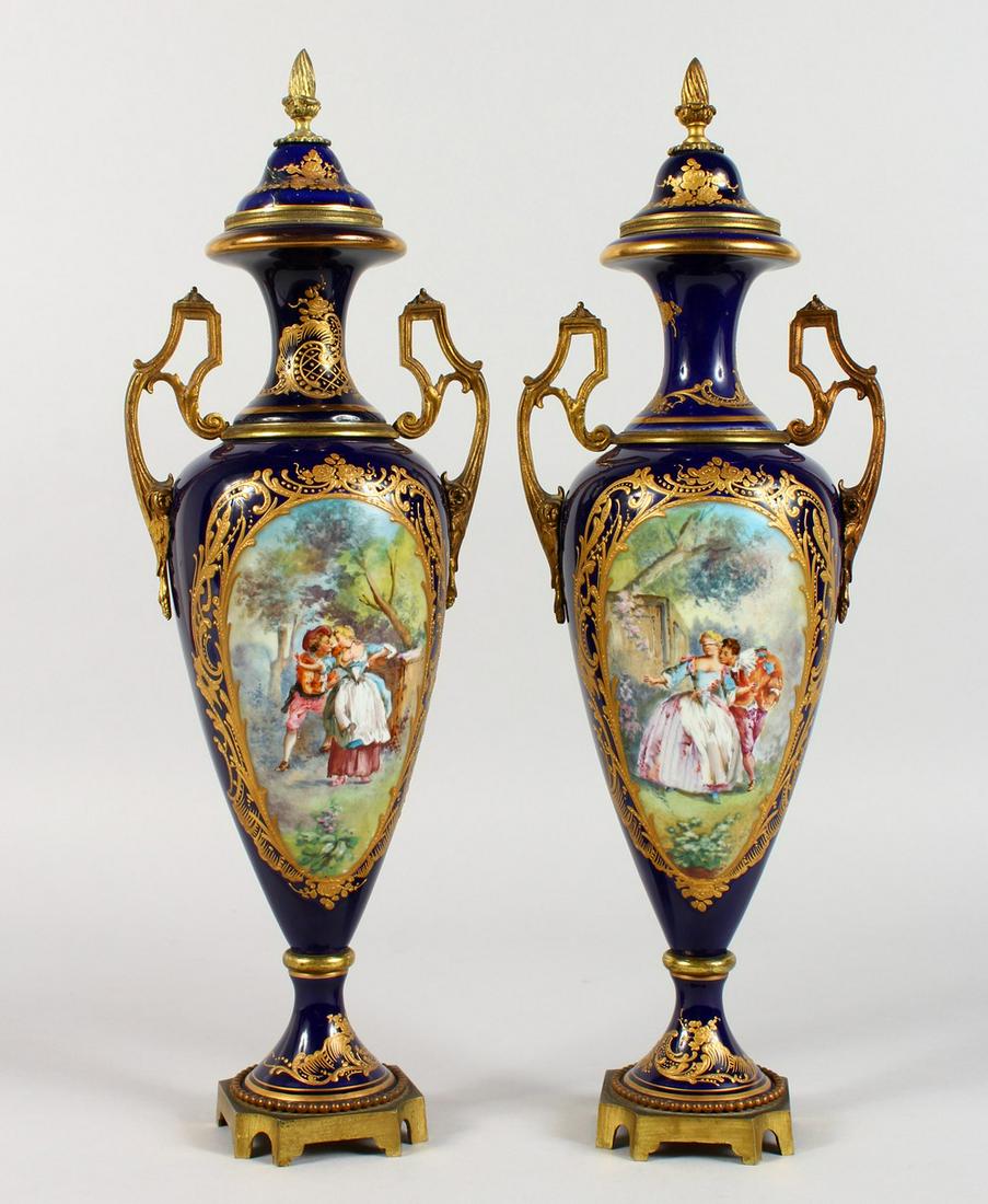 A SUPERB PAIR OF DARK BLUE GROUND SEVRES TWO-HANDLED: A SUPERB PAIR OF DARK BLUE GROUND SEVRES TWO-HANDLED URNS AND COVERS, with gilt decoration and ormolu mounts, the bodies painted with reverse panels of young lovers and a lake scene. 16ins high.